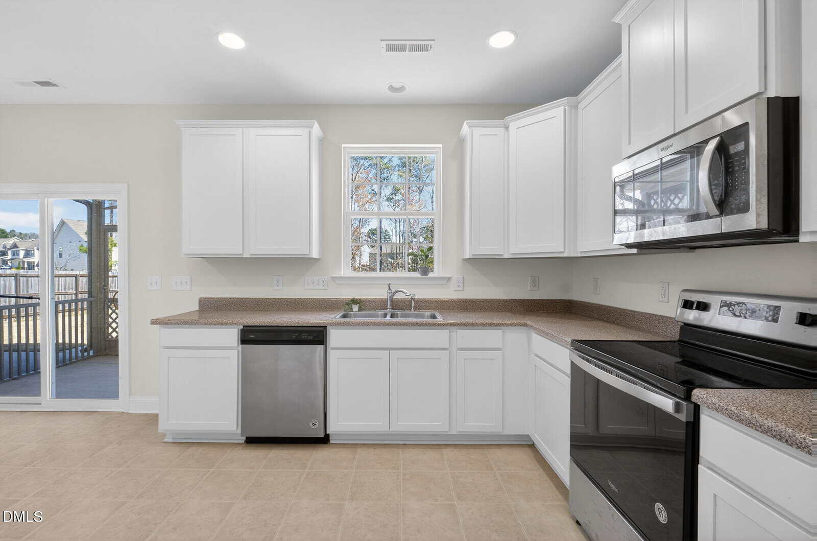 Modern kitchen with white cabinets, stainless steel appliances including dishwasher and microwave, double sink under a window, neutral tile floor, and a view of a deck and yard through a glass door on the left.