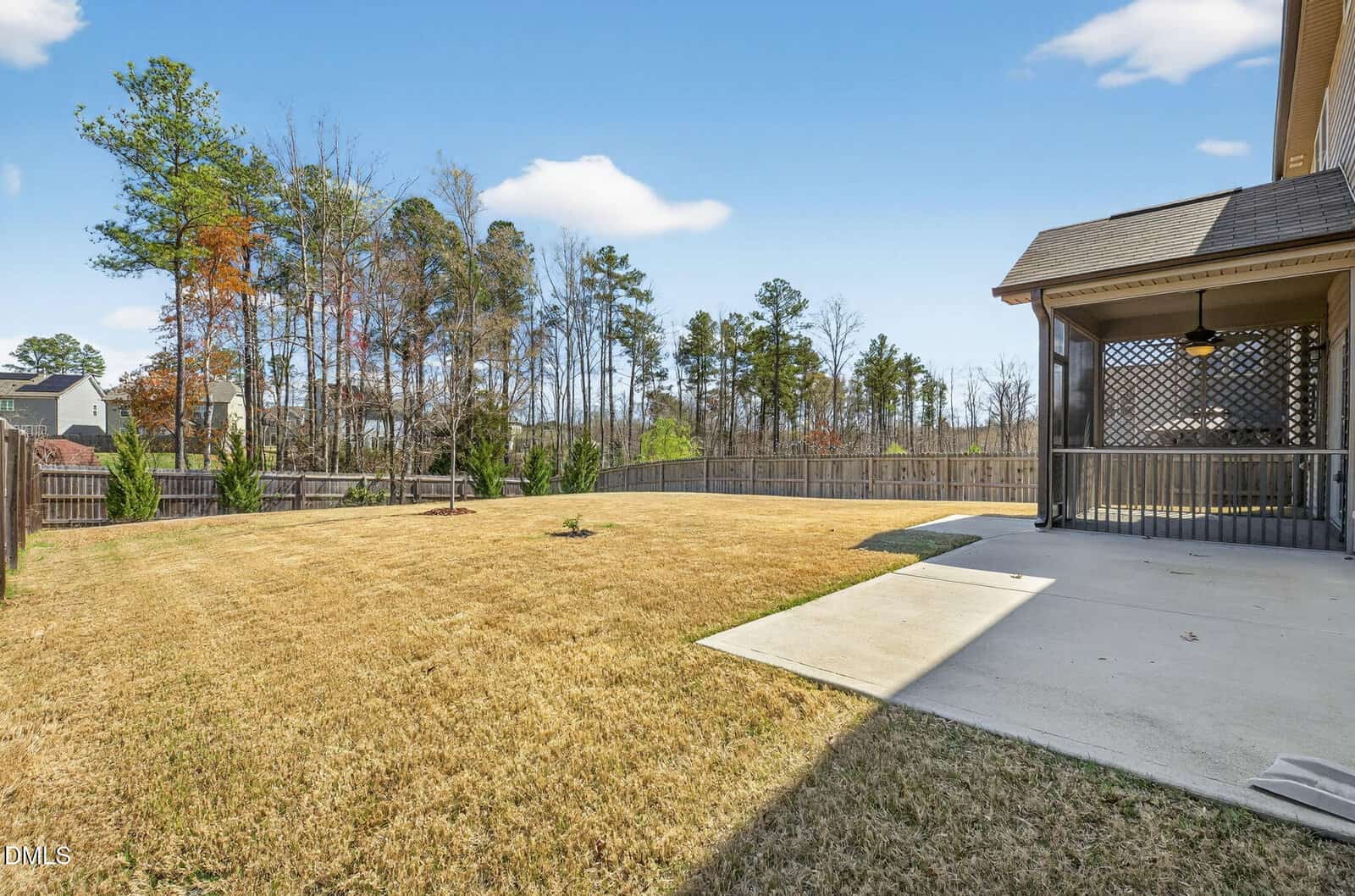 A spacious backyard with a large lawn, wooden fence, and a covered patio with a ceiling fan attached to a house. Trees are visible beyond the fence under a clear, blue sky.