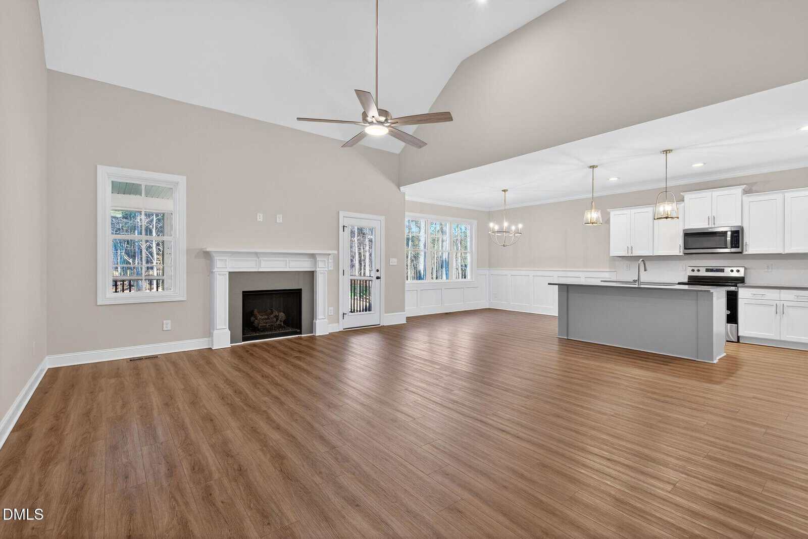 Spacious open-concept living area with wood floors, a ceiling fan, white fireplace, large windows, and an adjacent kitchen featuring white cabinets, stainless steel appliances, and a large island with pendant lighting.
