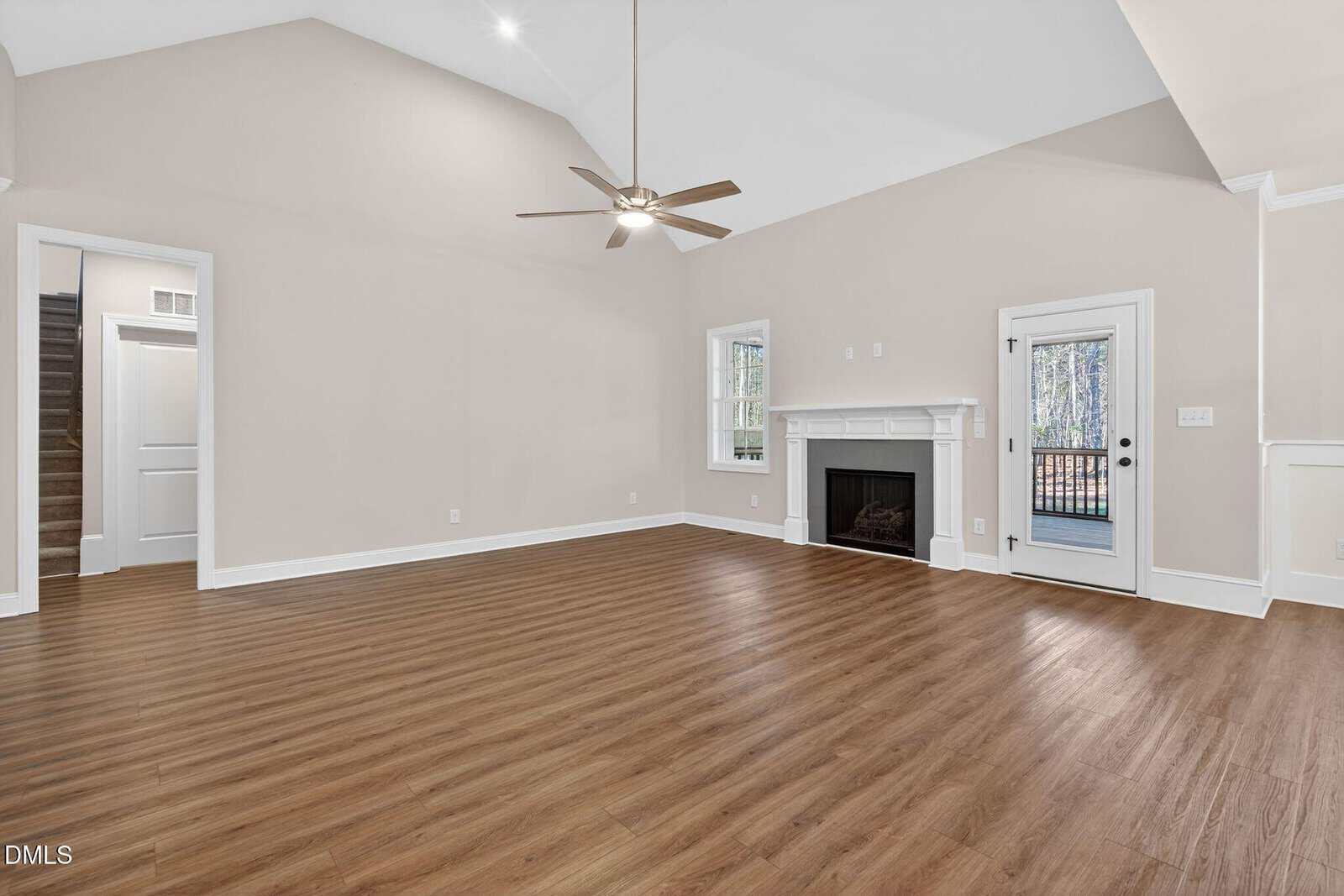 Spacious empty living room with wood flooring, beige walls, a ceiling fan, a fireplace, and a door leading to an outdoor deck. White trim and molding accent the room.