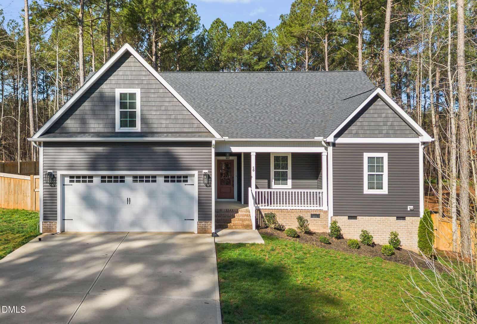 A single-story house with gray siding, white trim, and a two-car garage sits on a concrete driveway. There is a small front porch with railings, landscaped bushes, and trees in the background.