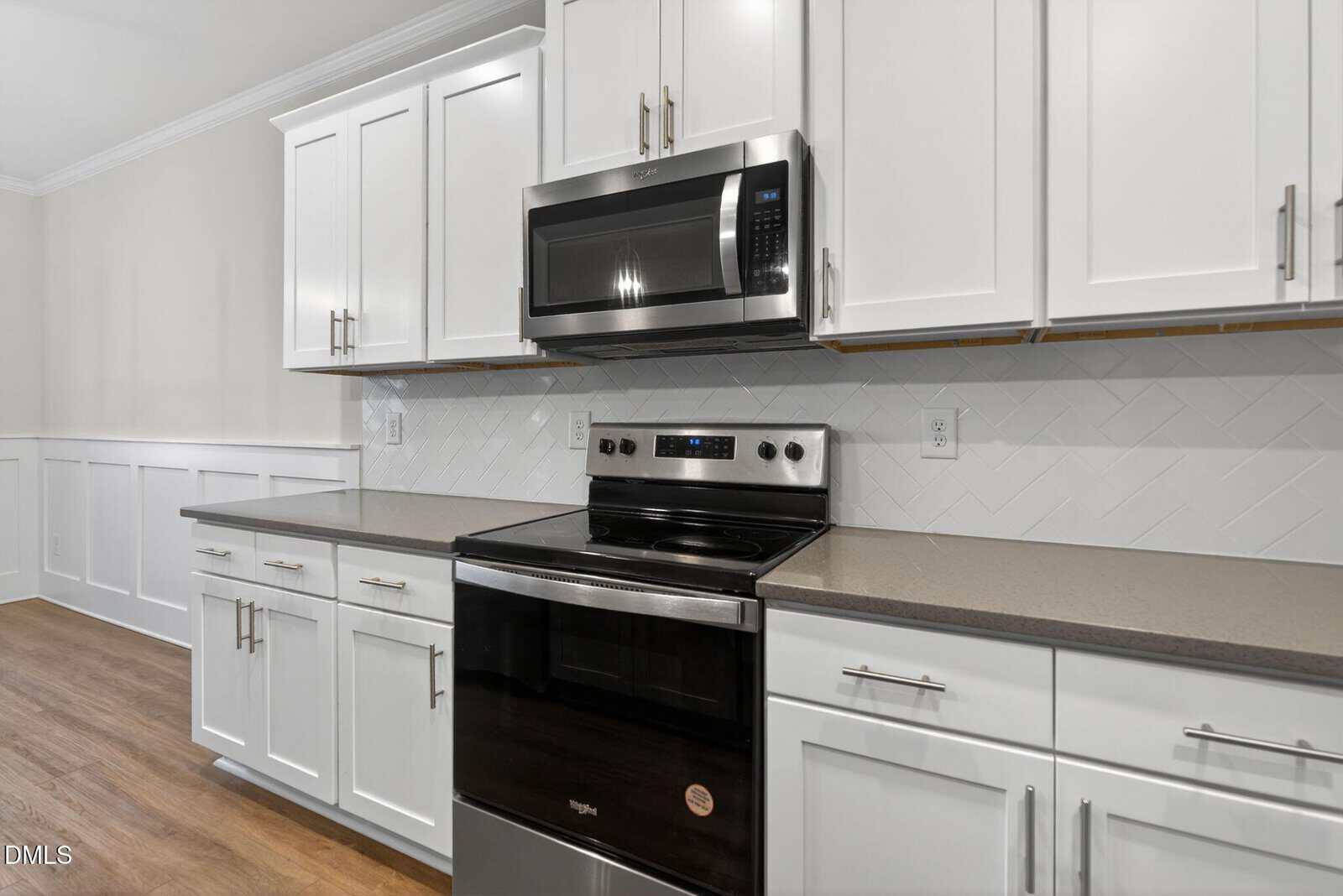 Modern kitchen with white cabinets, stainless steel microwave and oven, gray countertops, herringbone tile backsplash, and wooden floor. The space appears clean and well-lit, with simple, contemporary design elements.