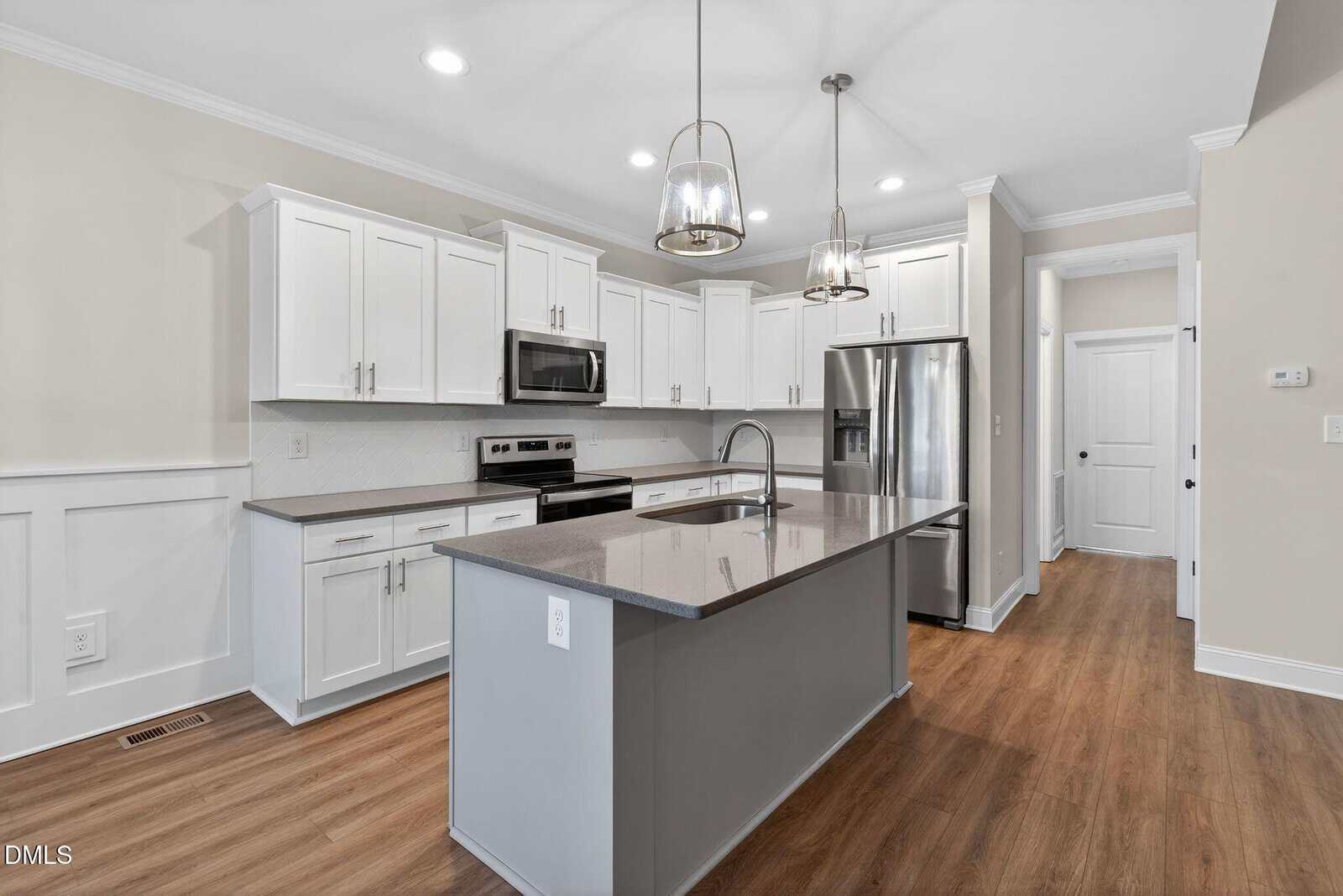 Modern kitchen with white cabinets, stainless steel appliances, gray island with sink, pendant lights, light walls, and wood flooring. The layout is open with recessed ceiling lights and a door in the background.