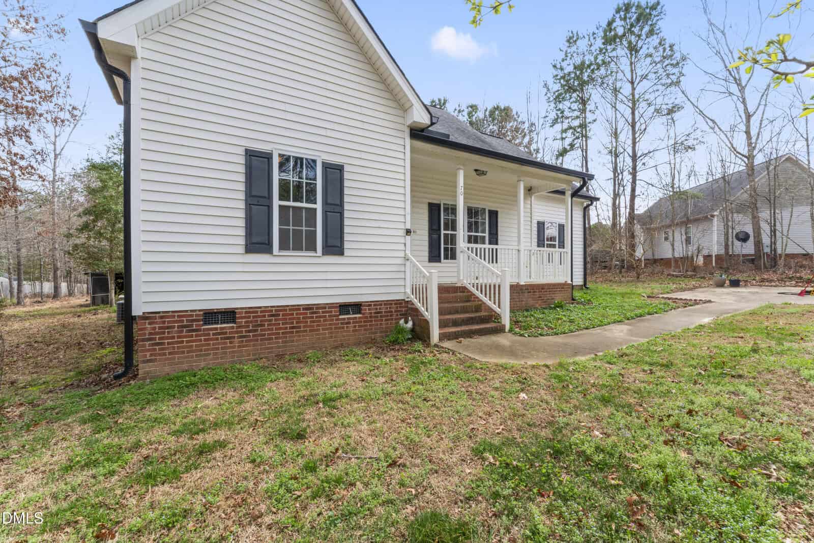 A white single-story house with black shutters, a brick foundation, and a covered front porch with white railings. A concrete walkway leads to the porch, and the yard is partly grass with patches of bare soil.