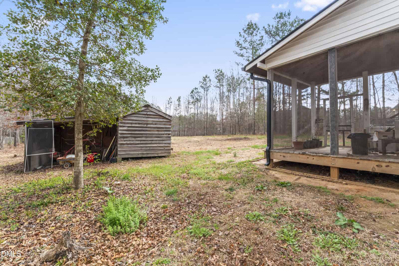 A screened porch with outdoor furniture is attached to a house on the right, and two wooden sheds are visible to the left, all surrounded by trees and a grassy, leaf-covered yard.