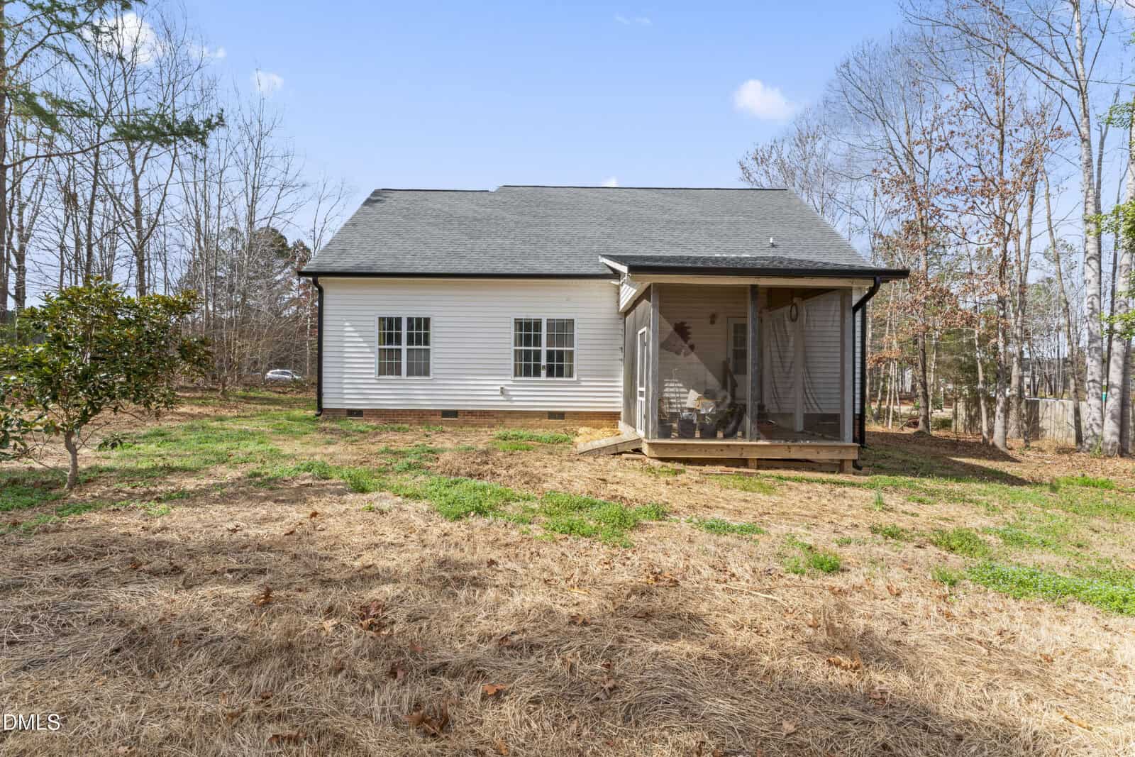A single-story white house with a gray roof is shown from the backyard. There is a small screened-in porch with steps leading to a grassy and mulched lawn, surrounded by trees.