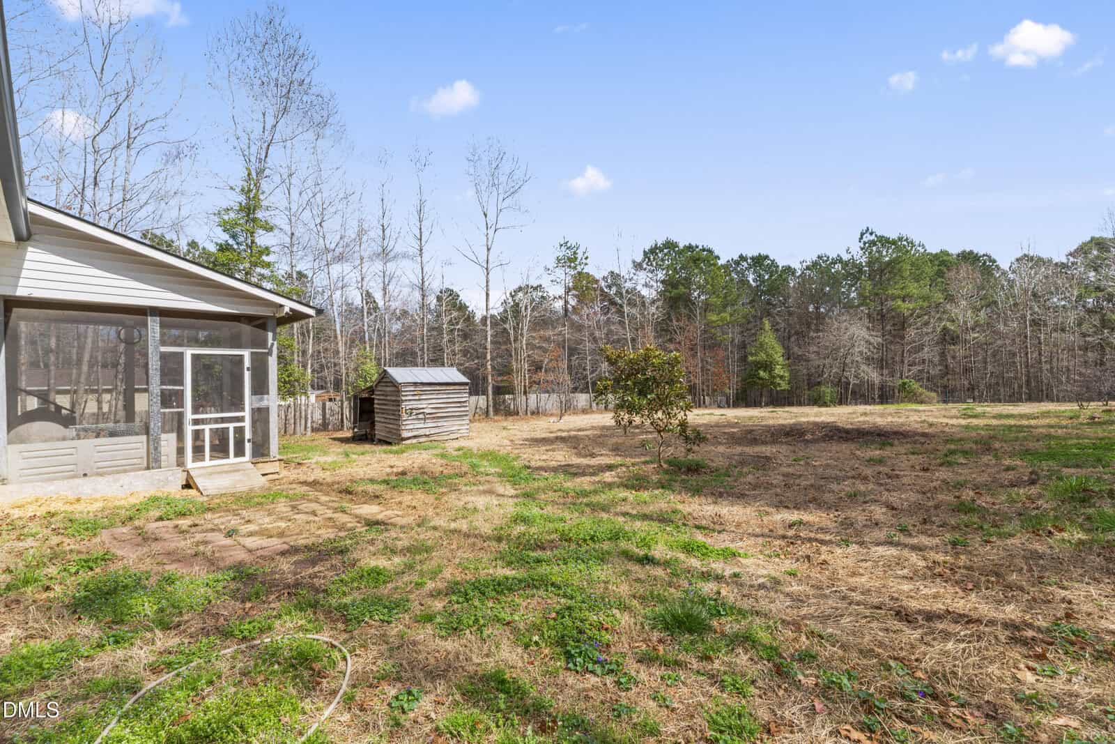 A backyard with sparse grass, a screened porch attached to the house, a small wooden shed, and a few trees. The area extends into a wooded line under a clear blue sky.