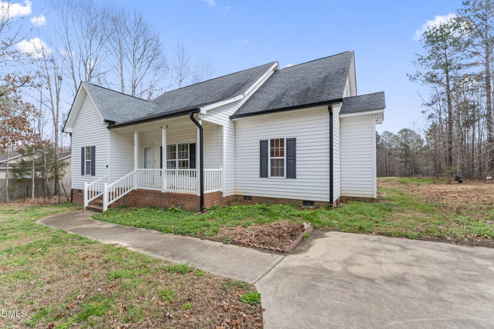 Single-story white house with black shutters and a covered front porch, set on a grassy lot with a concrete driveway and surrounded by trees under a clear blue sky.