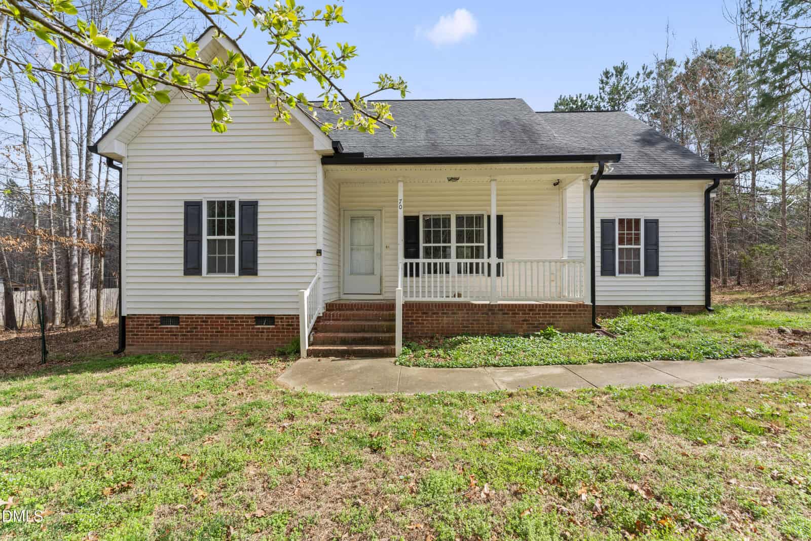 A single-story white house with black shutters, a covered front porch with white railings, brick foundation, and a concrete walkway, surrounded by grass and trees.