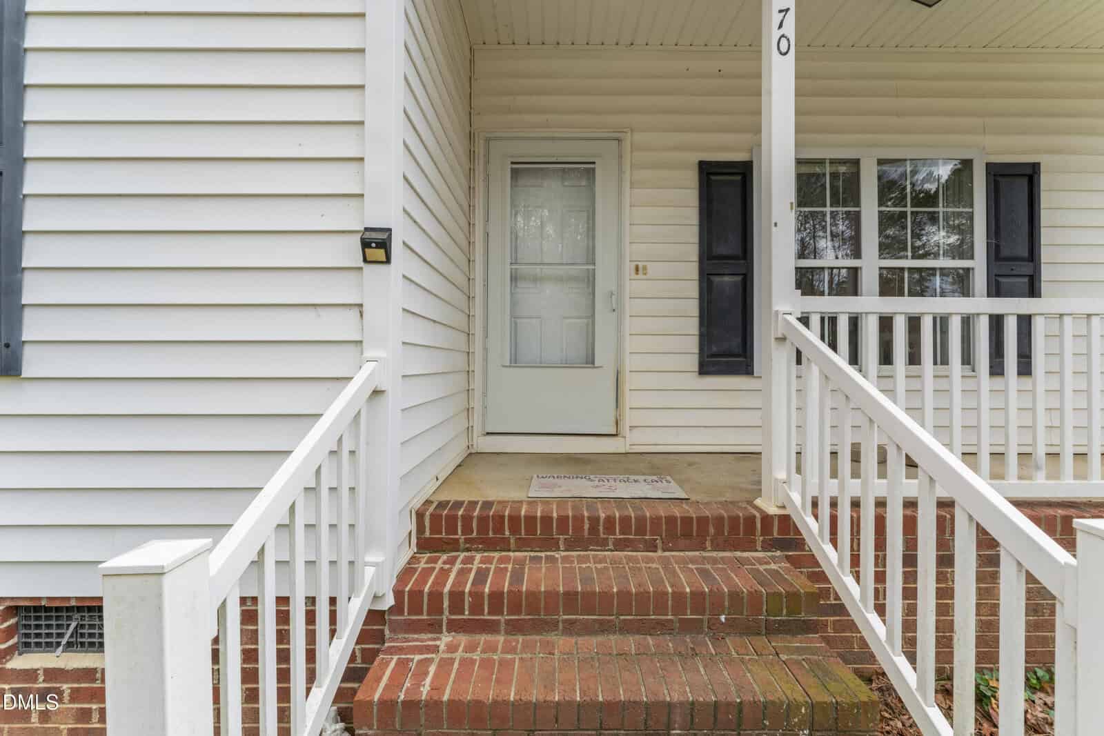 A brick staircase with white railings leads to a covered porch and a white front door with a glass storm door. The house has white siding, black shutters, and the number 70 visible above the entrance.