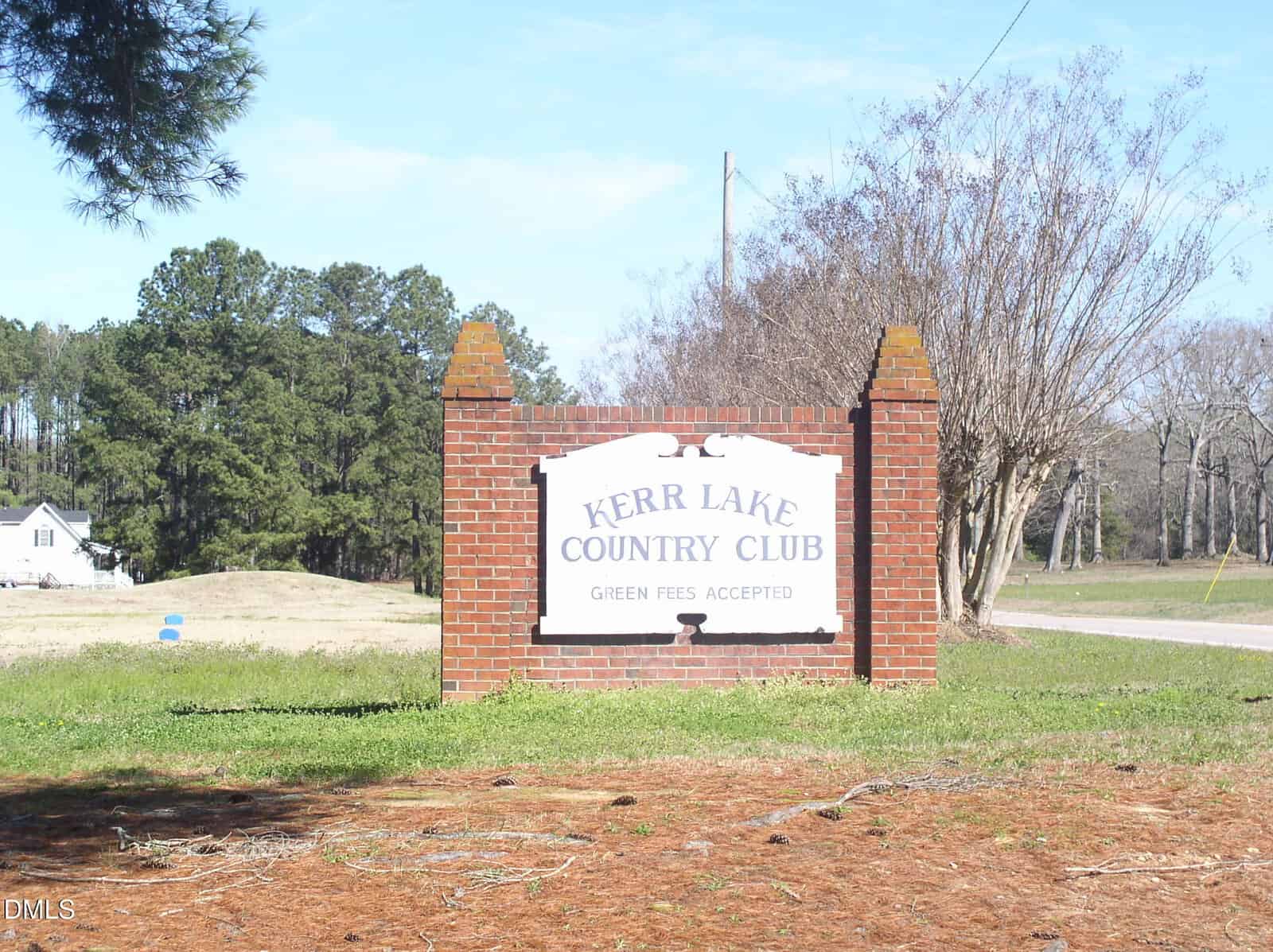 A brick sign at the entrance of Kerr Lake Country Club at 0 Country Lake Drive, Henderson, displays the club’s name and “Green Fees Accepted.” Trees, grass, and a house are visible in the background under a clear sky.
