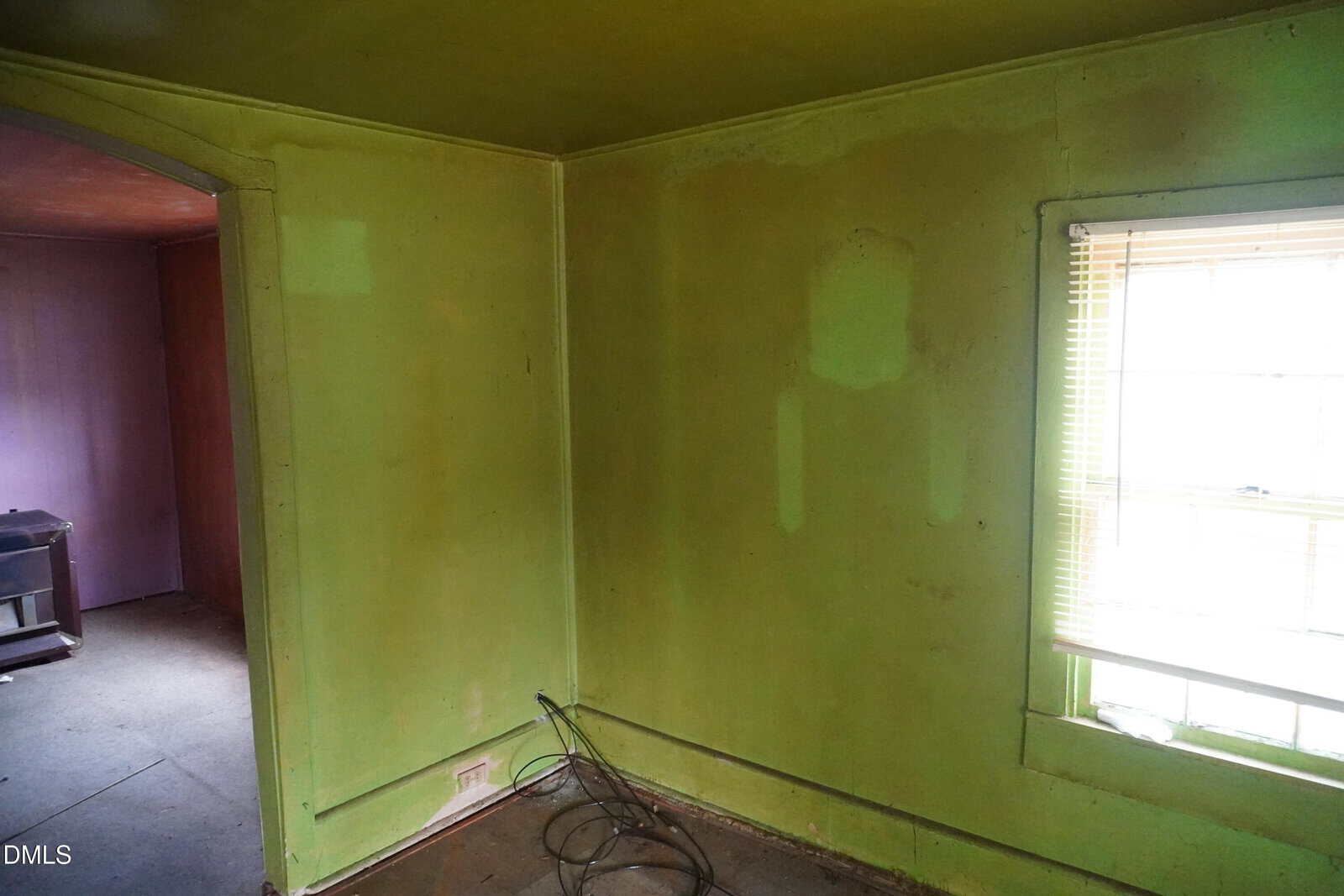 A small room with green walls, a window with blinds, some exposed cables on the floor, and a view into an adjacent purple-walled room. The room appears empty and in need of repairs or cleaning.