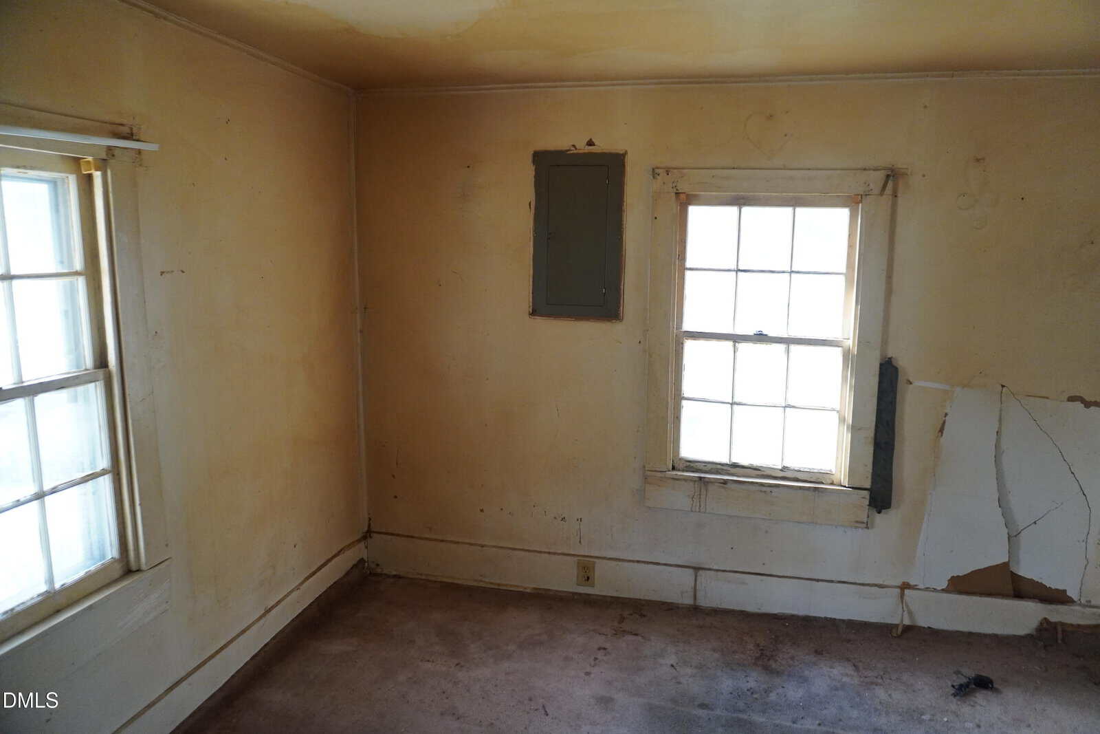 A small, empty room with stained, yellowed walls, two windows with peeling paint, a patch of cracked plaster, and a faded carpet. A gray electrical panel is mounted between the windows.