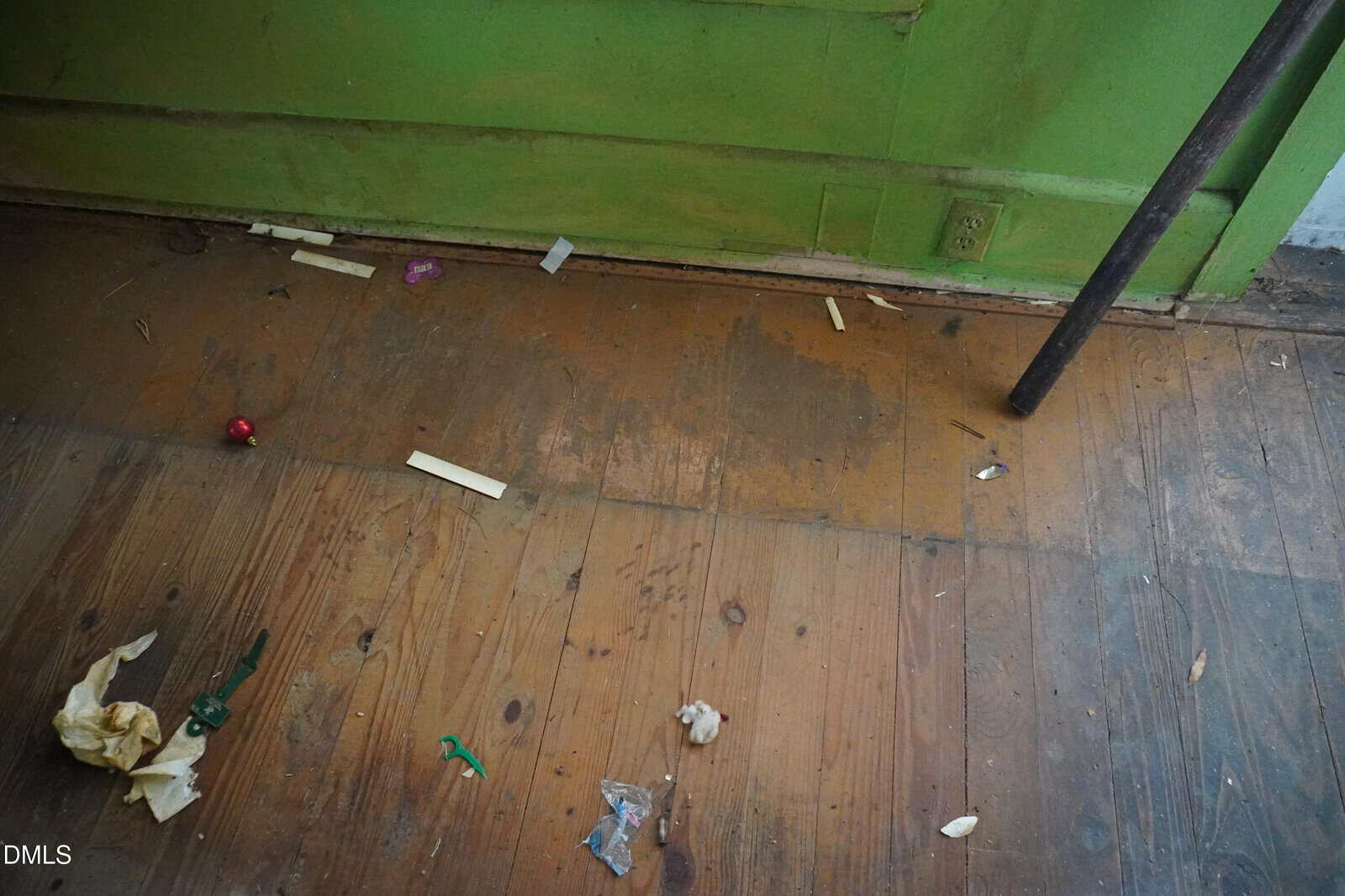 A worn wooden floor with scattered debris, including paper, plastic, and small objects, is shown near a green-painted wall with a power outlet and a metal pipe.