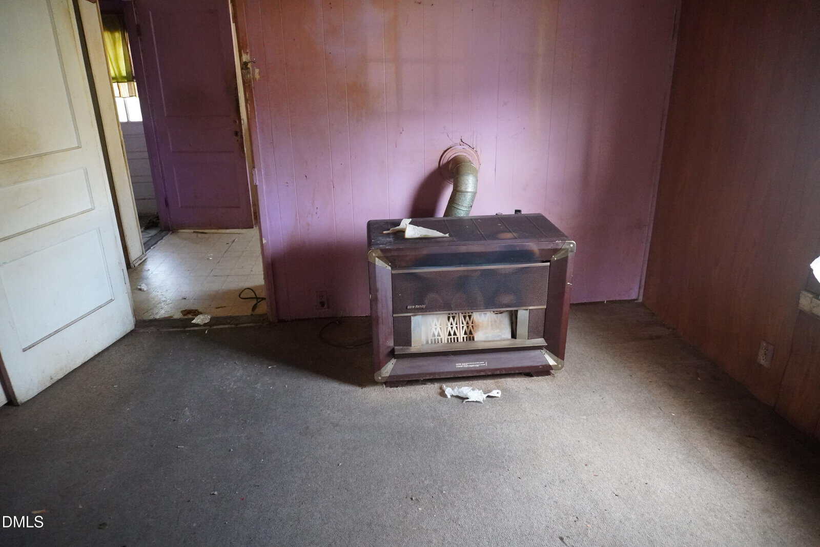 A small, old heating stove with a vent pipe is positioned against a purple wall in a mostly empty, carpeted room with worn surfaces. A paper towel rests on top of the stove. An open door leads to another room.