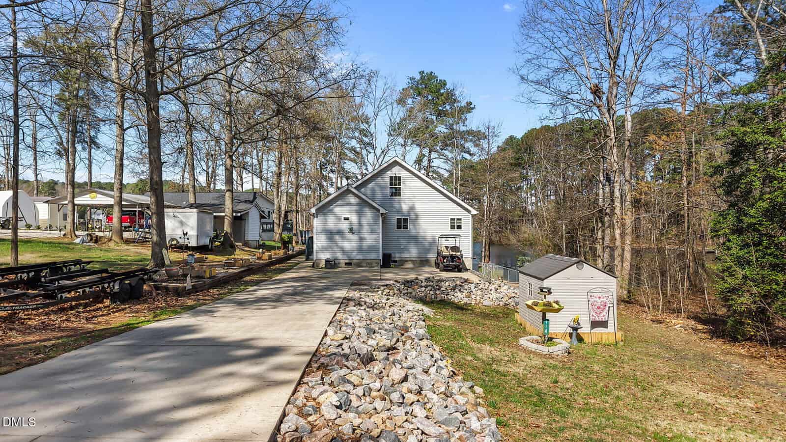 A light-gray house with a gable roof sits at the end of a concrete driveway, surrounded by trees. A small shed, birdbath, and rocks line the yard, and a lake is visible behind the house.