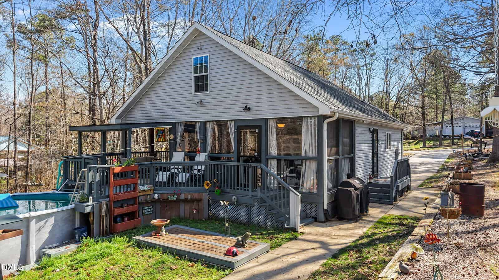 A small gray house with a screened porch, sloped roof, and side deck sits among leafless trees. There are plants, garden decorations, and outdoor furniture on the porch and yard. A paved driveway runs along the right side.