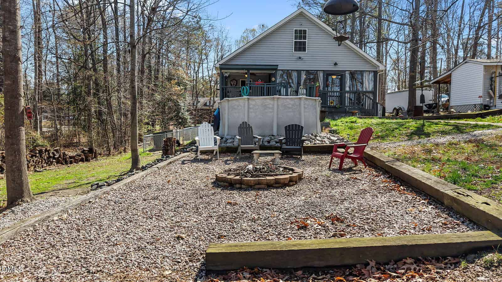 A backyard with a fire pit surrounded by four different-colored Adirondack chairs, bordered by wood beams and mulch, with a white house and trees in the background.