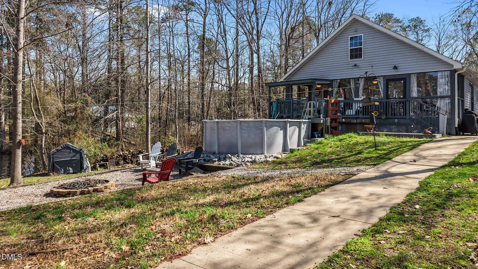 A gray house with a screened porch, raised pool, and wooden stairs sits on a grassy lot with a paved walkway. There is a fire pit area with red and brown chairs, and trees surround the property.
