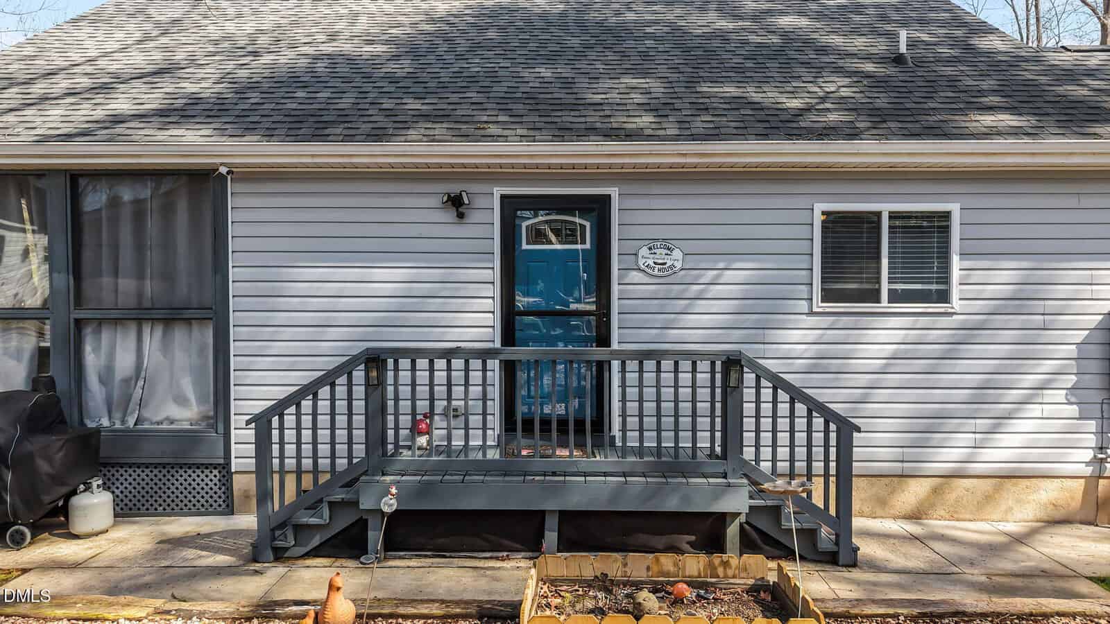 A small wooden porch with railings and steps leads to a blue door on a gray house. There is a barbecue grill on the left, a window on each side of the door, and a garden bed in the foreground.