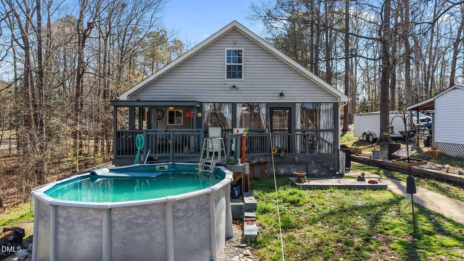 A small gray house with a screened porch and a wooden deck is shown. An above-ground pool is in the backyard, surrounded by grass and trees. A trailer and another house are visible to the right.
