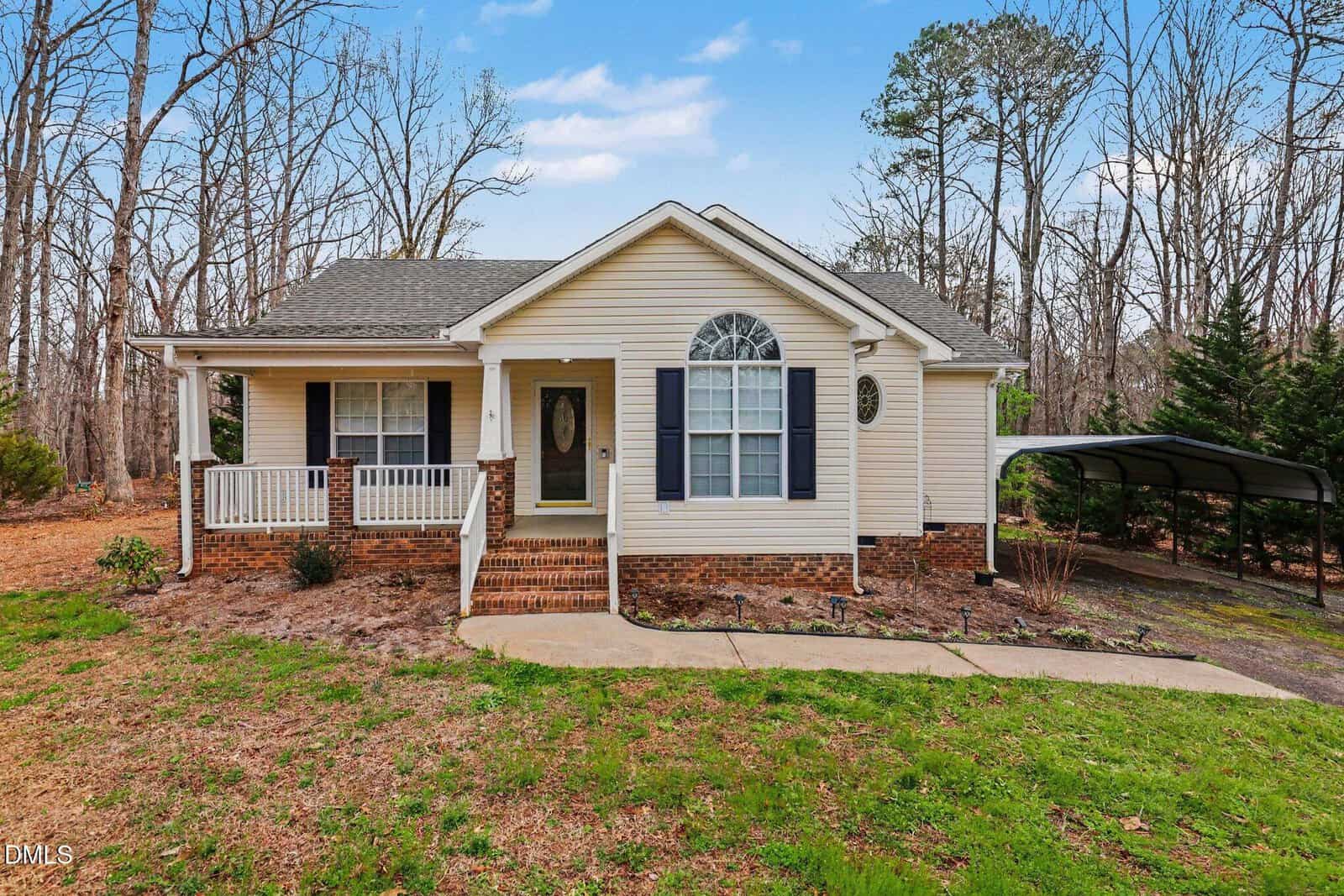 Single-story beige house with white trim, dark shutters, a covered front porch, brick foundation, and a carport on the right. The yard is grassy with a curved walkway and surrounded by bare trees.