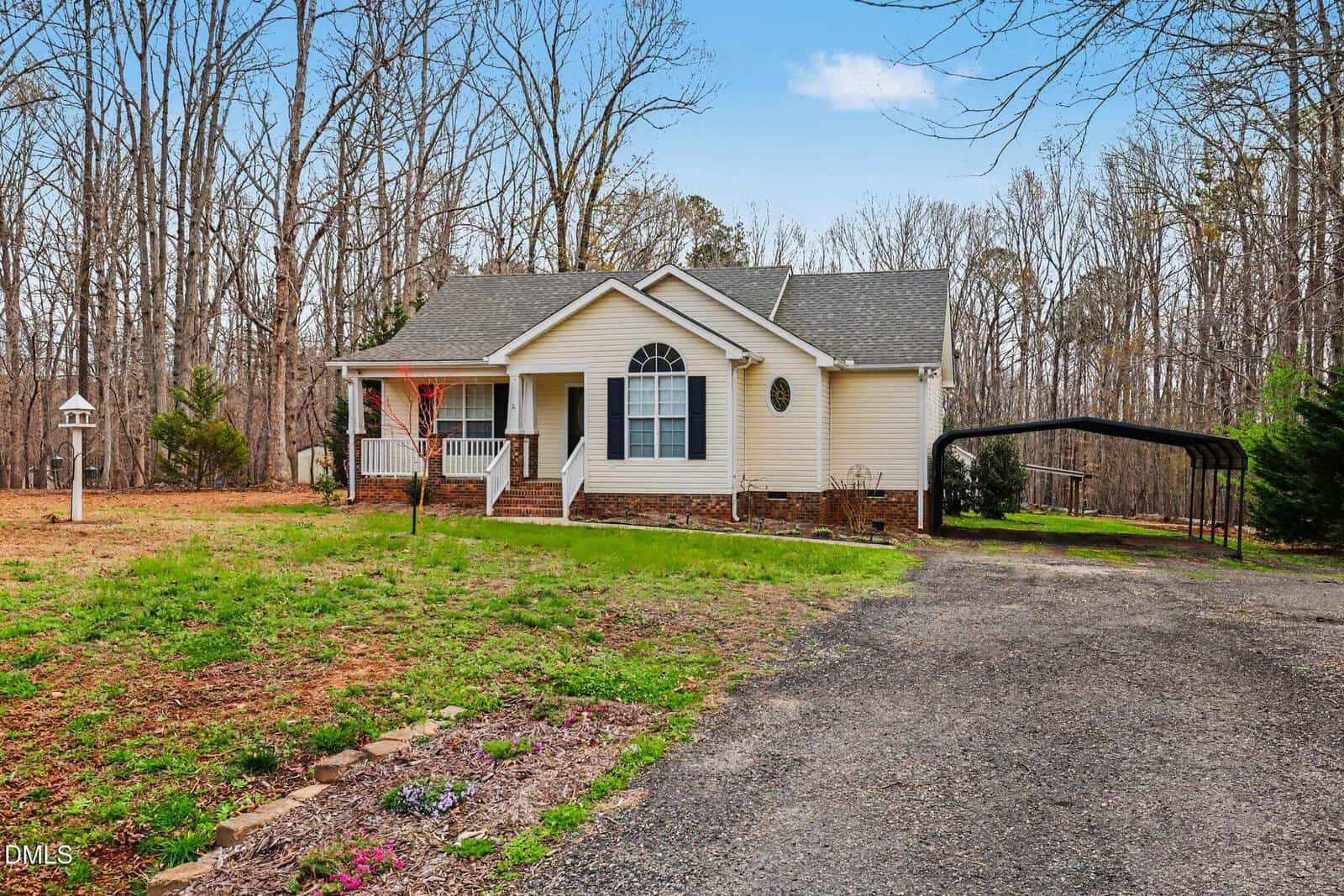 Single-story house with a covered front porch, white siding, and brick foundation, set on a large lot with a gravel driveway, carport, and surrounding trees without leaves.