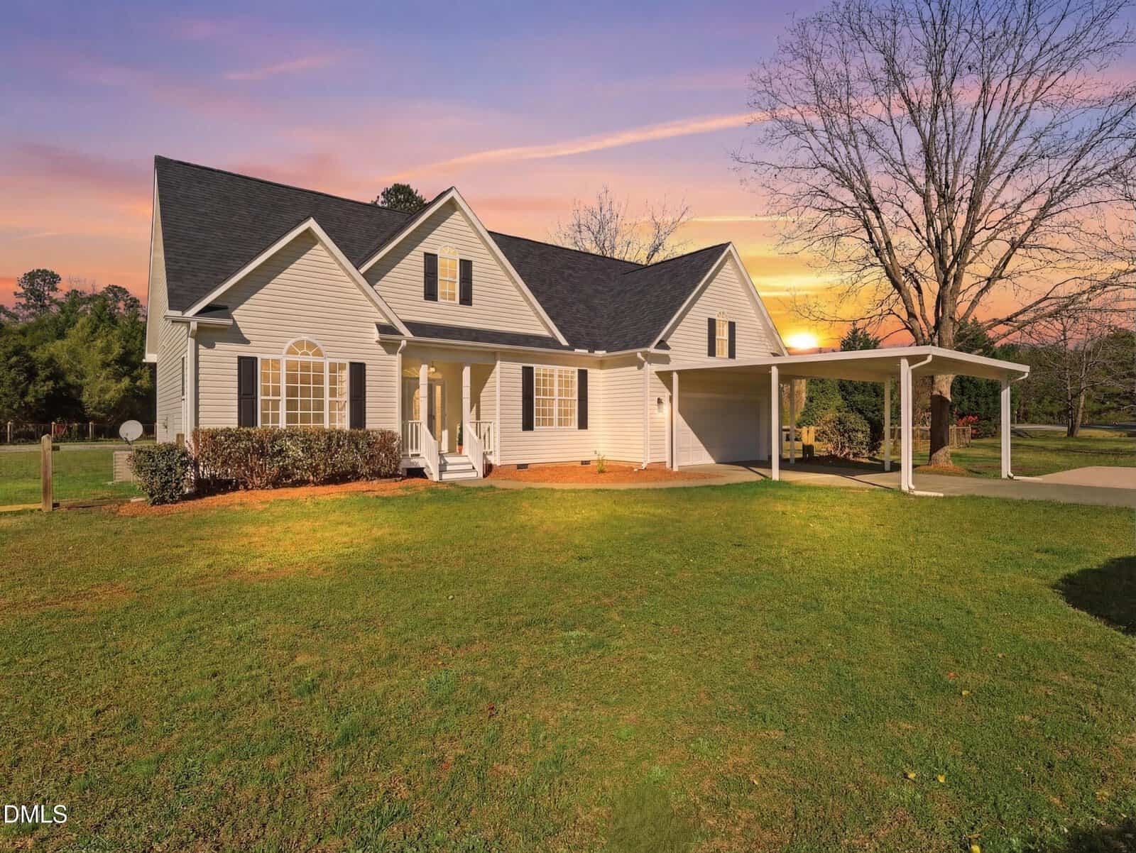 Single-story house with beige siding, black roof, covered front porch, and attached carport, set on a large green lawn at sunset with a bare tree to the right and shrubs along the front of the house.