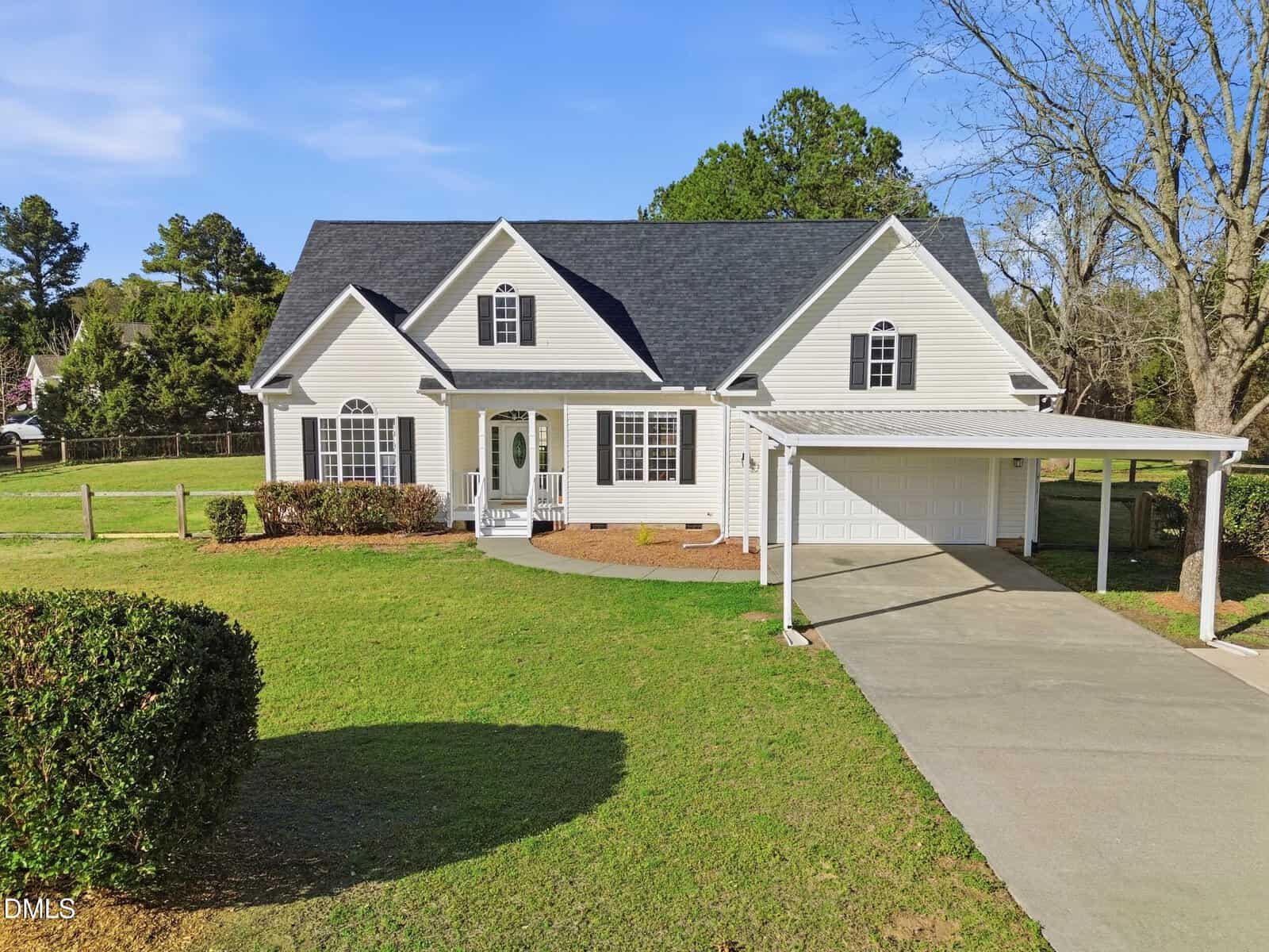 A two-story light-colored house with dark roofing, an attached two-car garage, and a covered carport. The property features a wide driveway, well-kept lawn, shrubs, and several mature trees in the background.