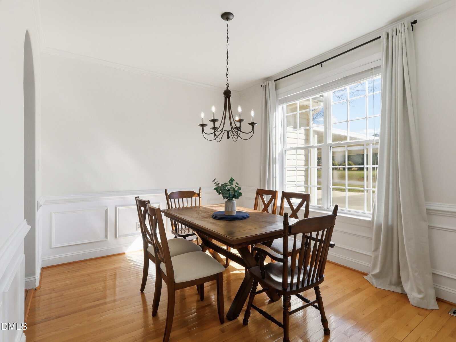 Dining room with a wooden table, six chairs, a black metal chandelier, white curtains, large window, white walls with molding, and hardwood floors. A small plant is centered on the table. Natural light enters the room.