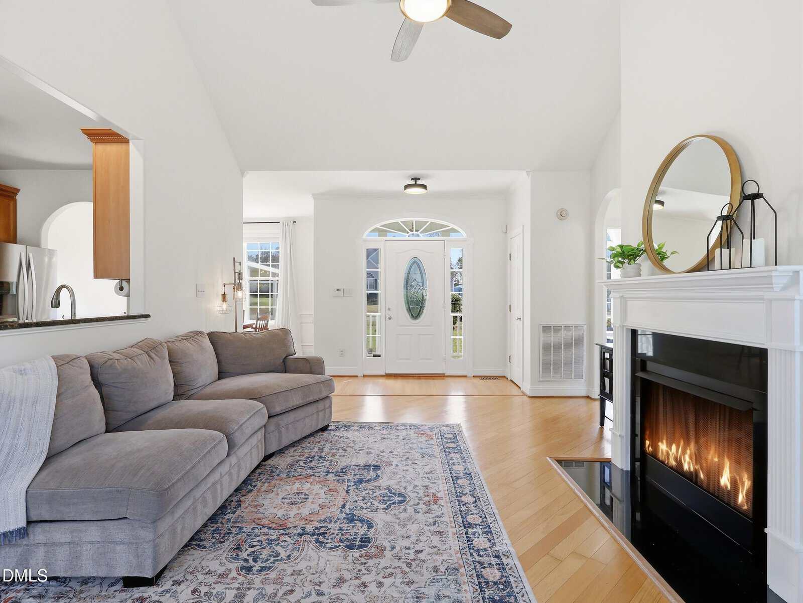 Bright living room with a gray sectional sofa, patterned rug, modern fireplace, and ceiling fan. The entrance door with glass panel is centered, and kitchen area is partially visible on the left. Walls and ceiling are white.