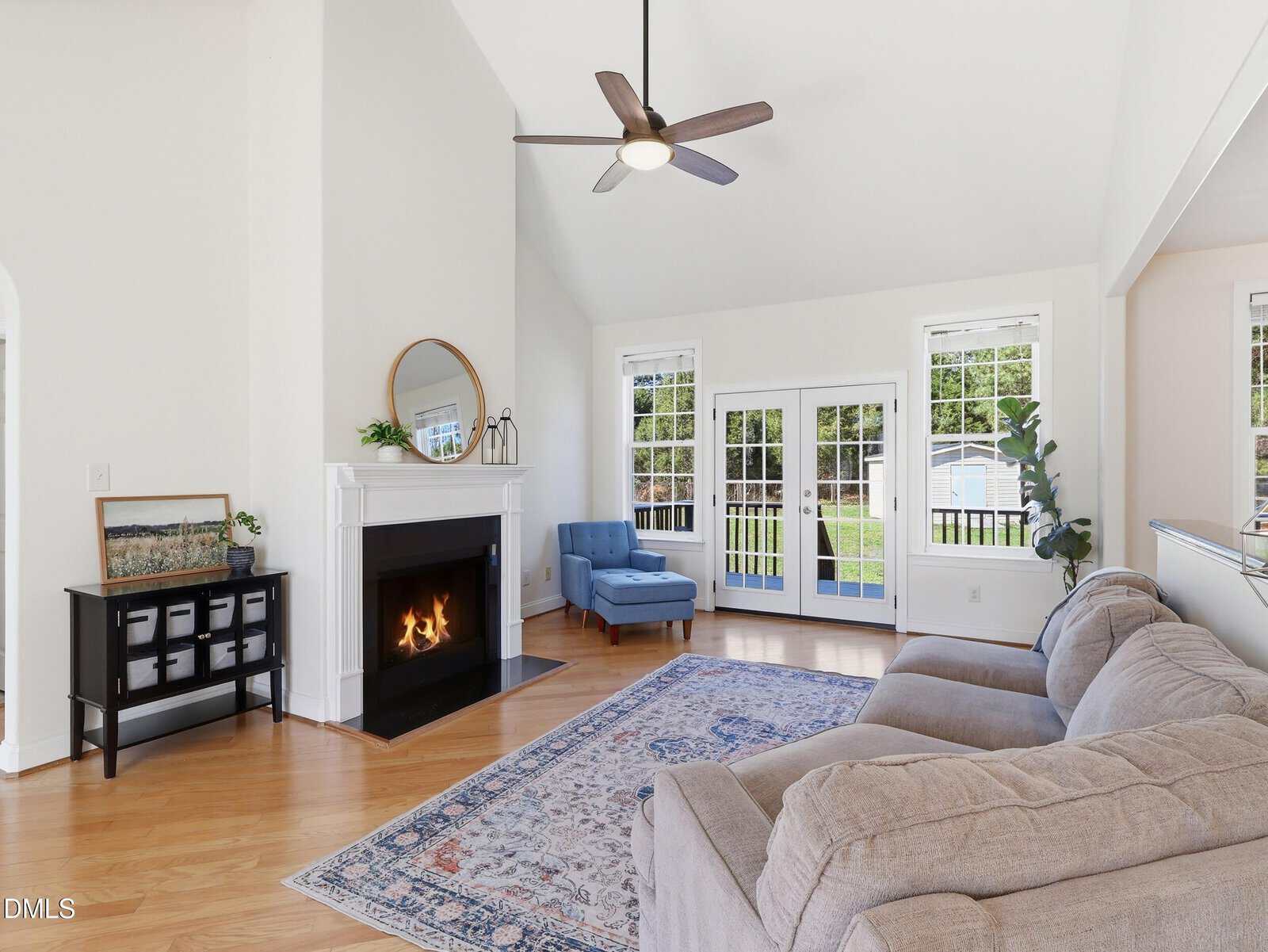 A spacious living room with a beige sectional sofa, a blue armchair by a window, a lit fireplace with a round mirror above, a patterned rug, and French doors leading outside. Ceiling fan above.
