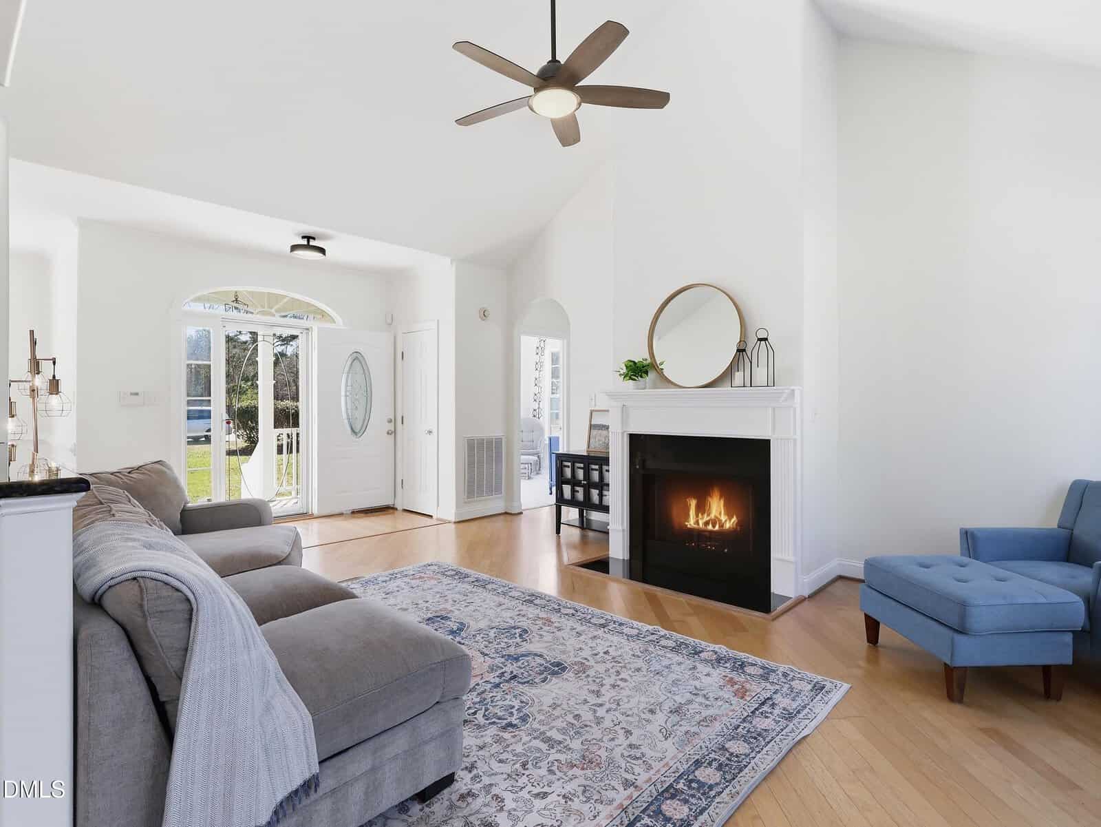 A bright living room with a gray sectional sofa, blue accent chair, patterned rug, and a lit fireplace beneath a round mirror. Light wood floors and a ceiling fan are visible, with sunlight coming through the front door.