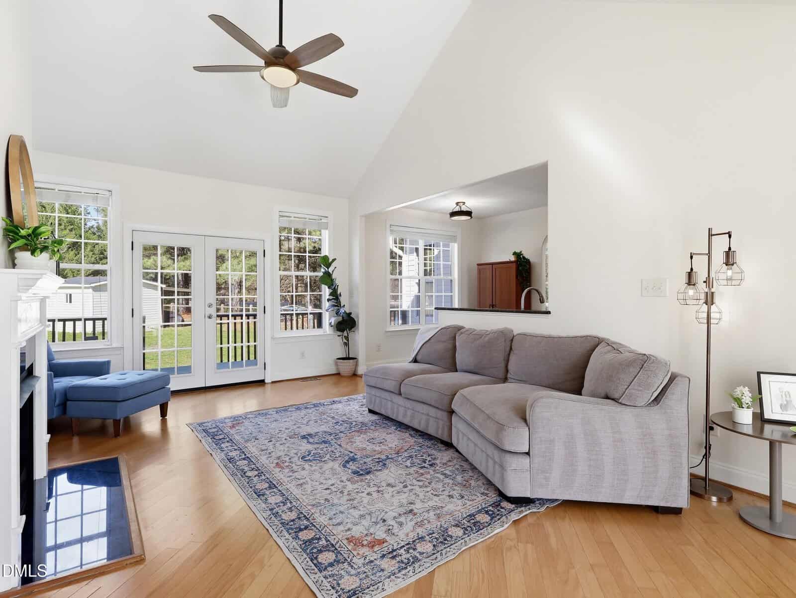 Bright living room with light wood floors, a gray sectional sofa, patterned area rug, ceiling fan, large windows, and glass doors leading outside. There’s a blue chaise by the windows and a glimpse of the kitchen beyond the sofa.