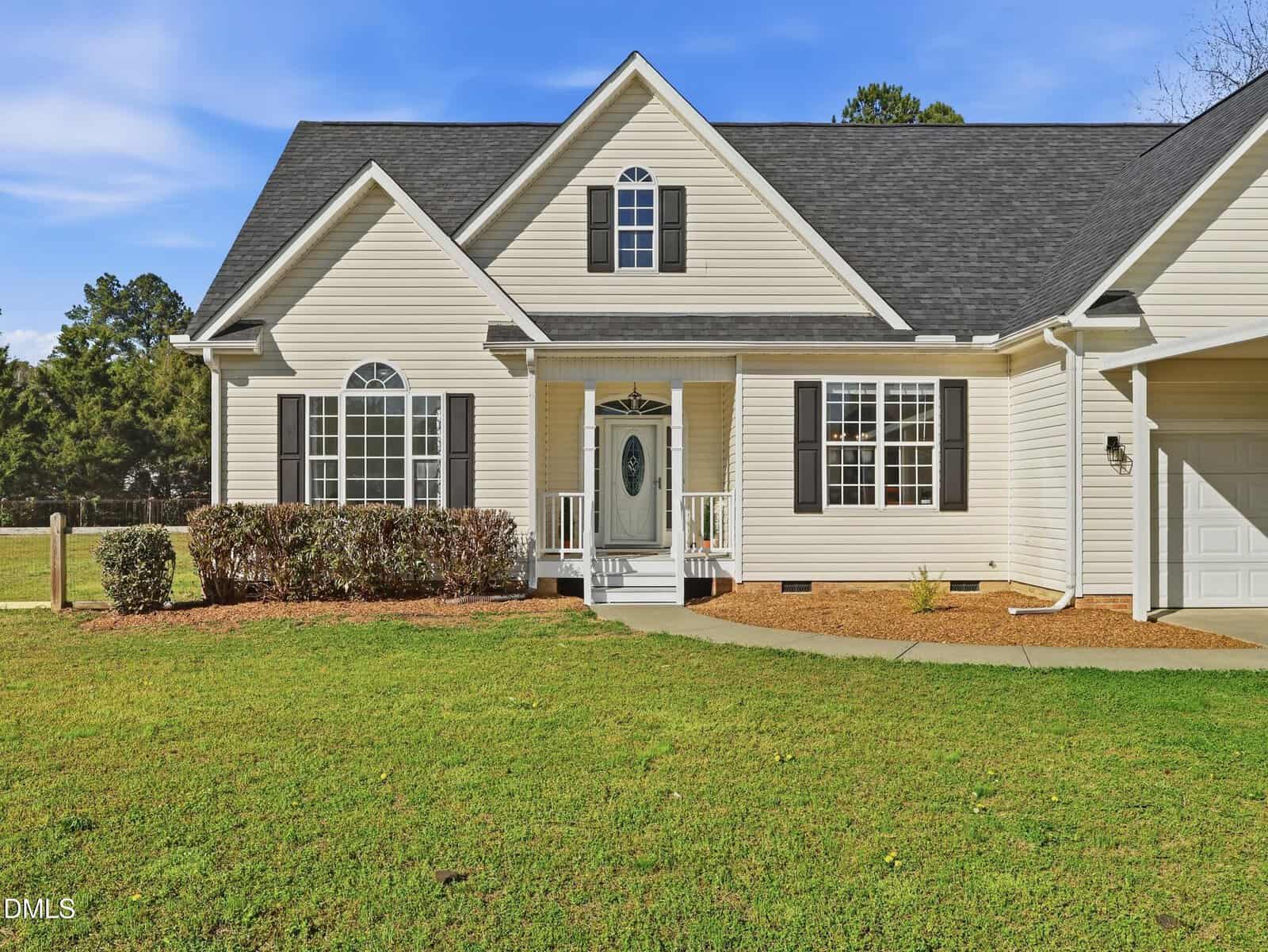 A beige single-story house with black shutters, a gabled roof, white trim, and a small front porch. The lawn is green and neatly maintained, with shrubs along the front and a driveway leading to a garage.