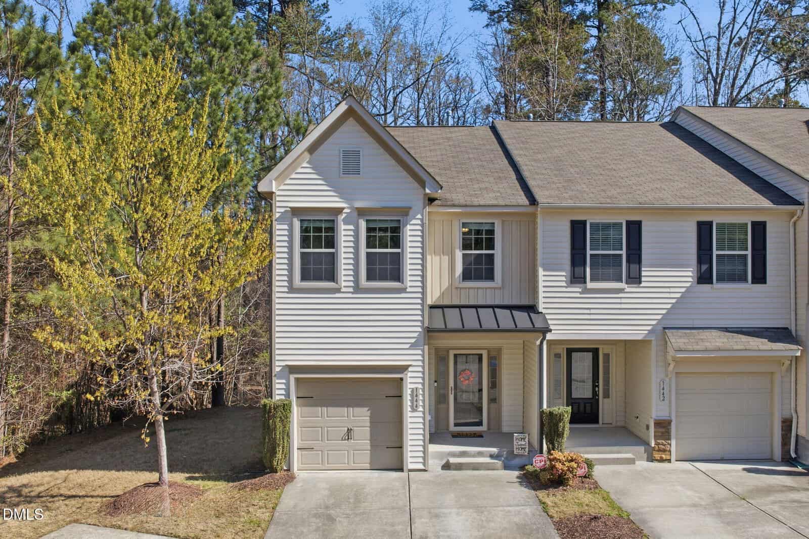 Two-story beige townhouse with attached single-car garage, black front door, and small covered porch. There is a yellow-leaved tree and a patch of grass on the left, with woods in the background.