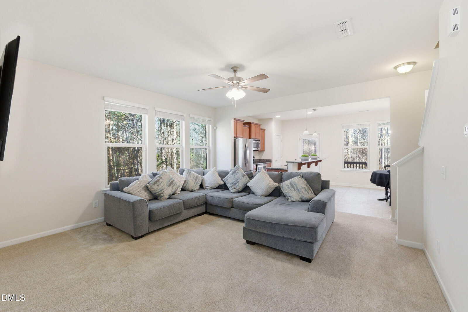 A spacious living room with beige carpet, a large gray sectional sofa with patterned pillows, ceiling fan, and three large windows. The area opens to a kitchen and dining space with hardwood flooring and stainless steel appliances.