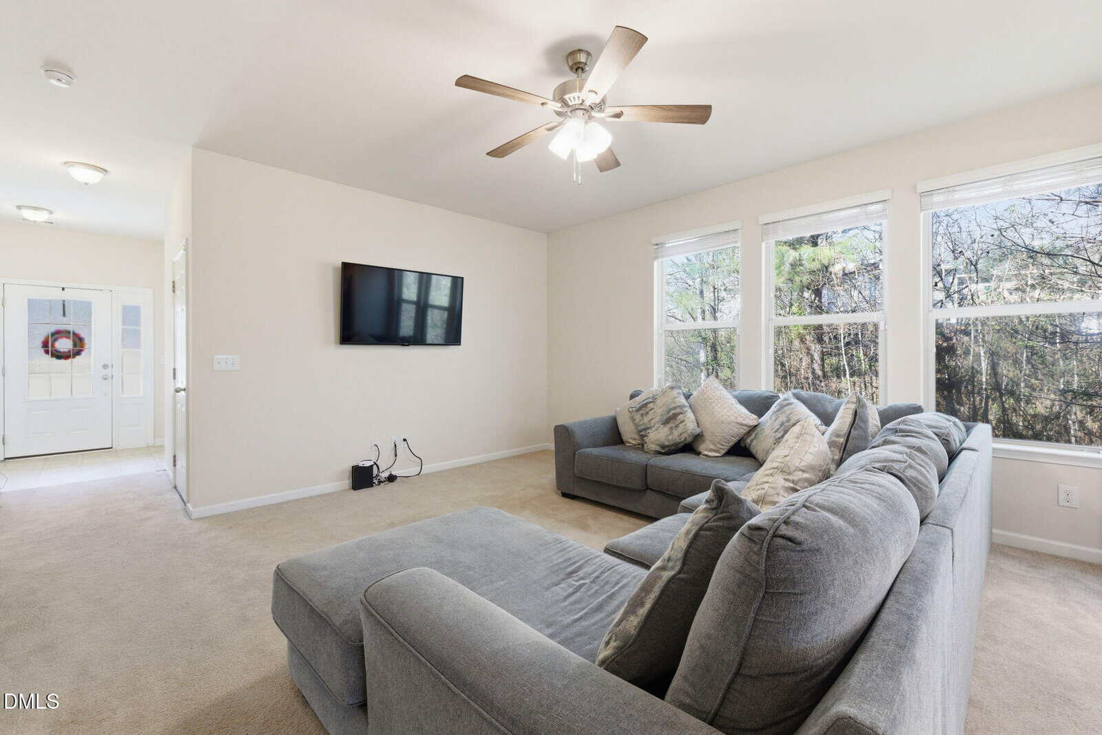 A living room with beige carpet, two gray sectional sofas, a wall-mounted TV, ceiling fan with light, and three large windows showing trees outside. Entryway with a wreath on the door is visible in the background.