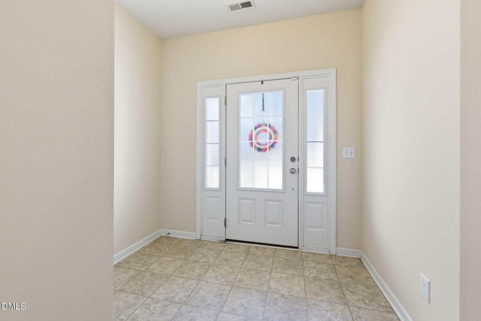 A simple entryway with beige walls, tiled floor, and a white front door with glass panels. A red and blue wreath hangs on the door. Natural light enters through the windows.