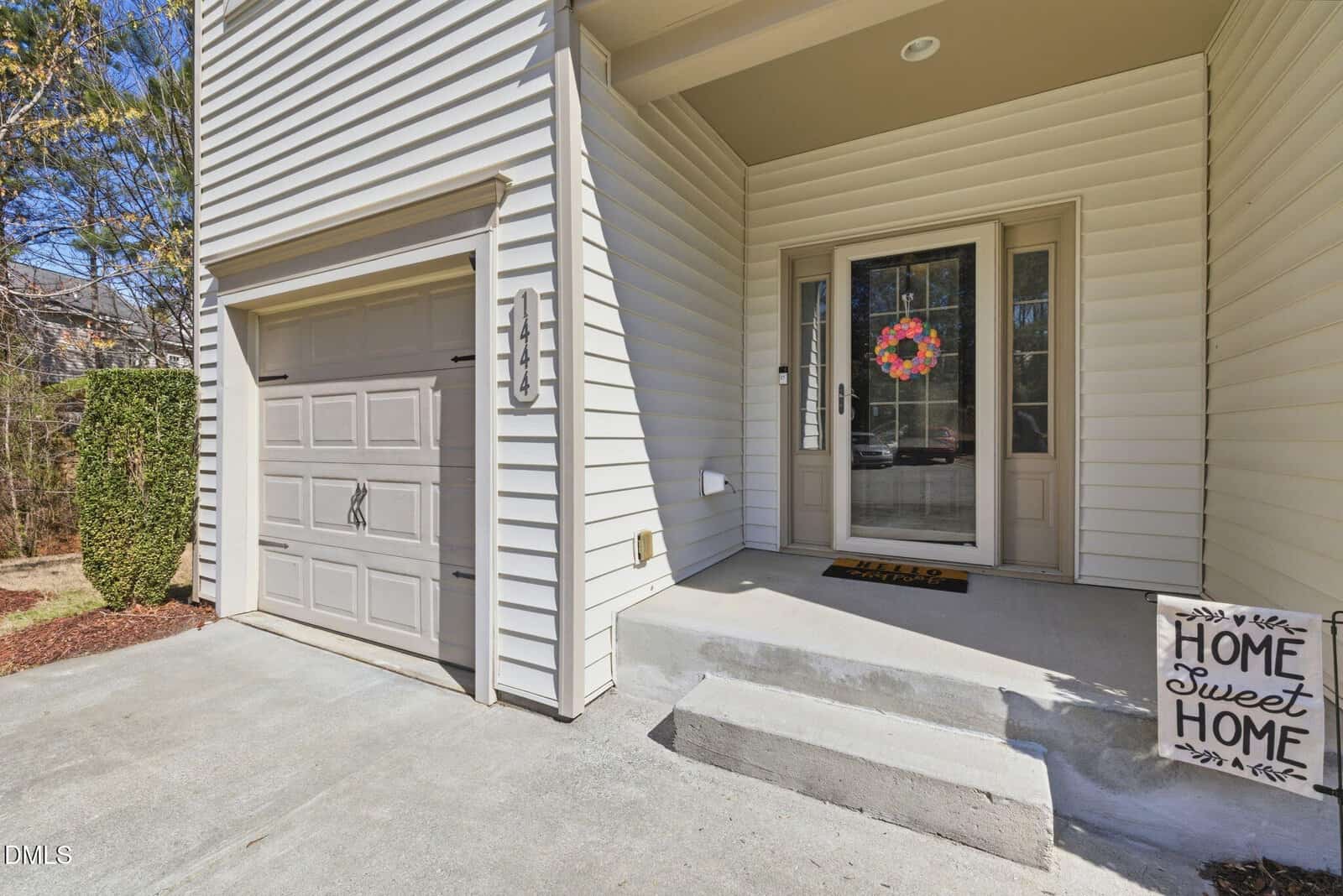 A beige house exterior with a garage door on the left, a front door with a floral wreath in the center, and a doormat and sign reading "Home Sweet Home" on the concrete steps. Shrubs and trees are visible in the background.