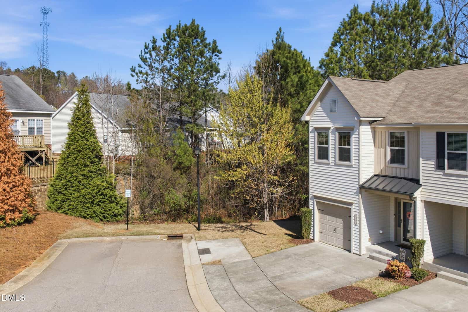 A two-story beige townhouse with a single-car garage and small front porch sits at the end of a cul-de-sac, surrounded by trees and neighboring houses under a clear blue sky.
