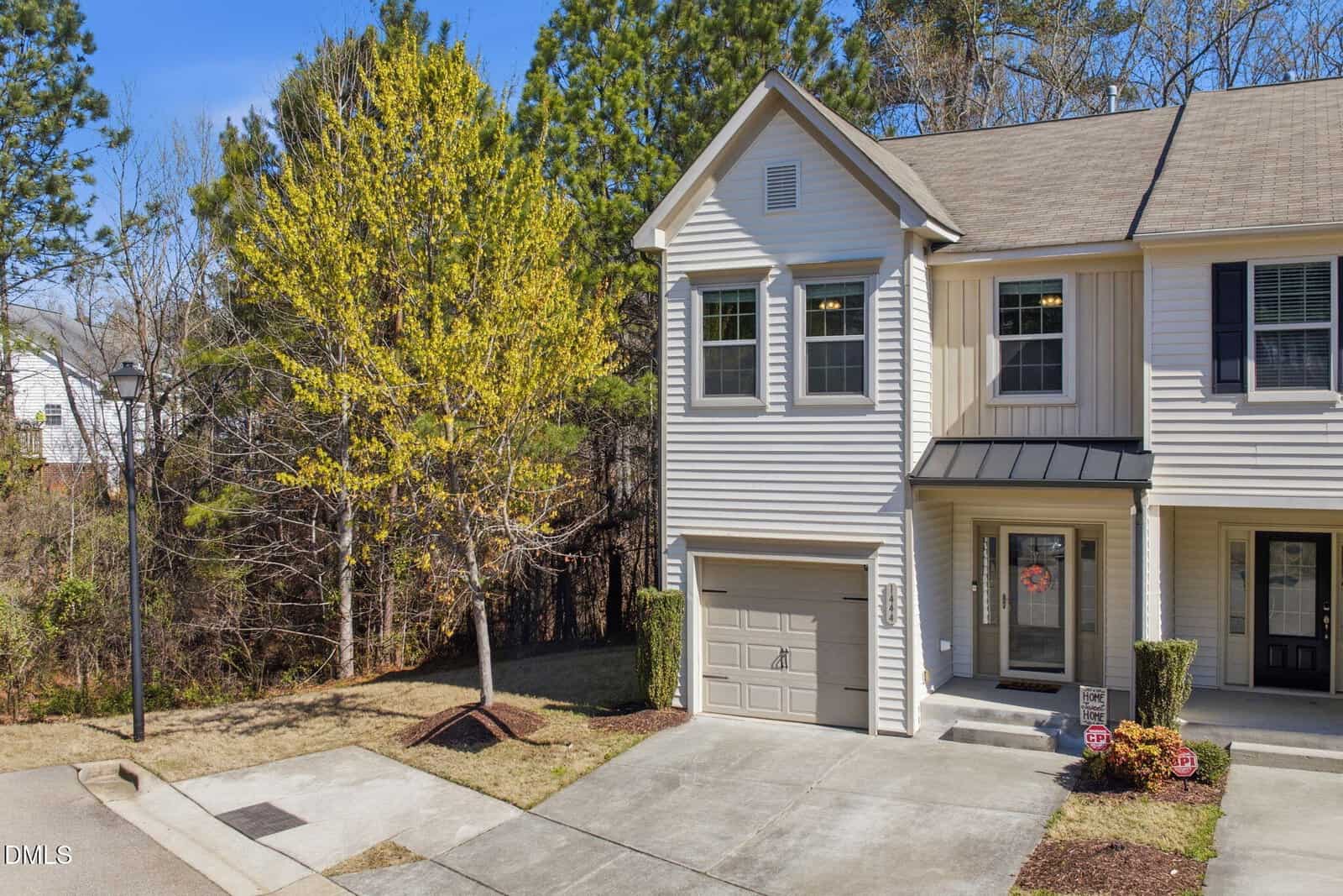 A two-story beige townhouse with a single-car garage and a small covered entryway, neighboring another unit, sits next to a tree and a wooded area under a clear blue sky.