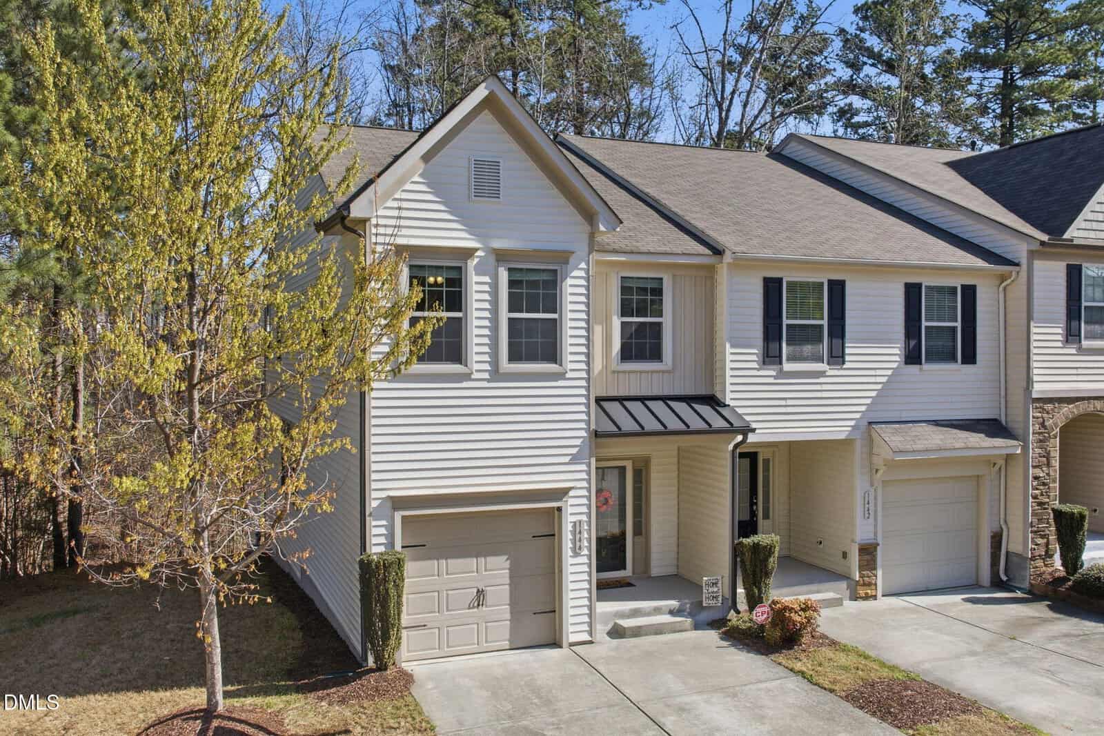 Two-story beige townhouse with a one-car garage and covered front porch, located at the end of a row. There's a small front yard with a tree and shrubs, and the driveway leads to the garage. Tall trees are visible in the background.