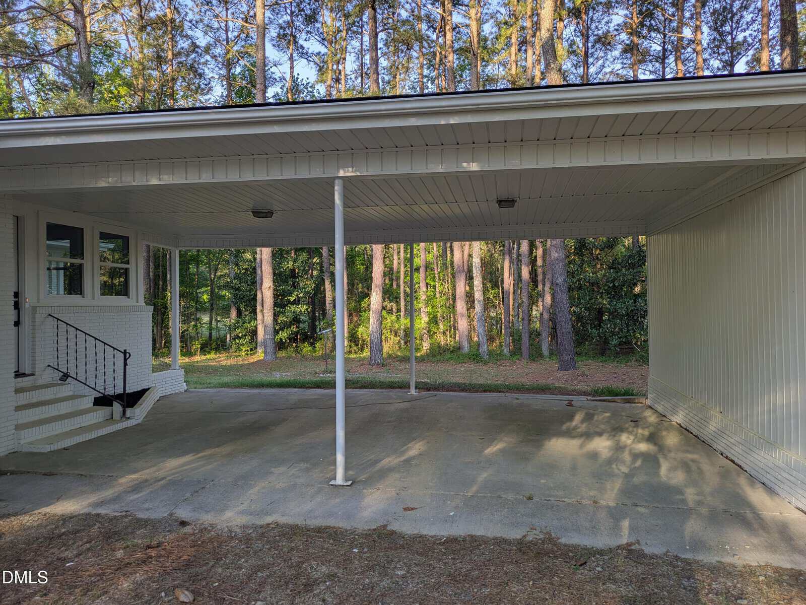 A covered carport with white columns, a concrete floor, and steps leading to a white brick house. Pine trees and greenery are visible in the background.