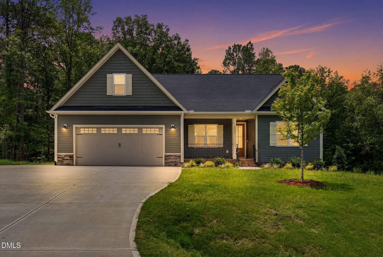 A modern single-story house with a two-car garage, front porch, and well-lit exterior, set against a sunset sky and surrounded by lush trees and lawn.