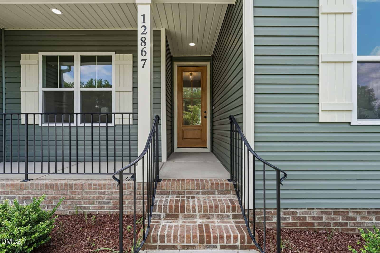 Front entrance of a house with green siding, white trim, brown brick steps, black metal railings, and a wooden front door with glass panel. House number 12867 is displayed vertically by the door.
