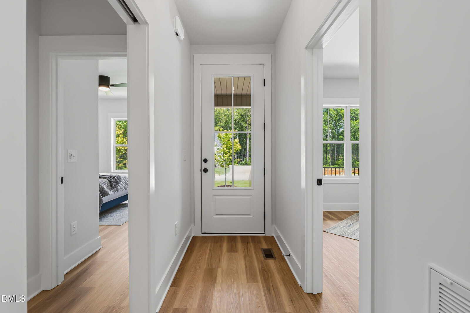 Bright hallway with light wood floors, white walls, and doors leading to bedrooms. A glass door at the end opens to a yard with green trees visible outside. Natural light fills the space.