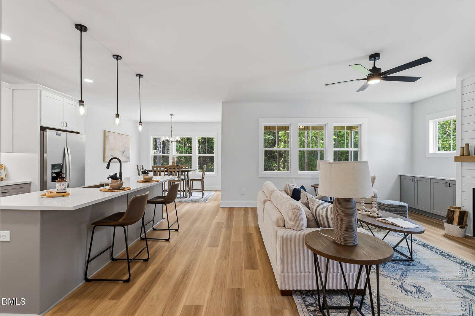 Open-concept kitchen and living room with light wood floors, white cabinets, pendant lights, a dining area, large windows, beige sectional sofa, ceiling fan, and a fireplace. Bright, modern, and spacious interior.