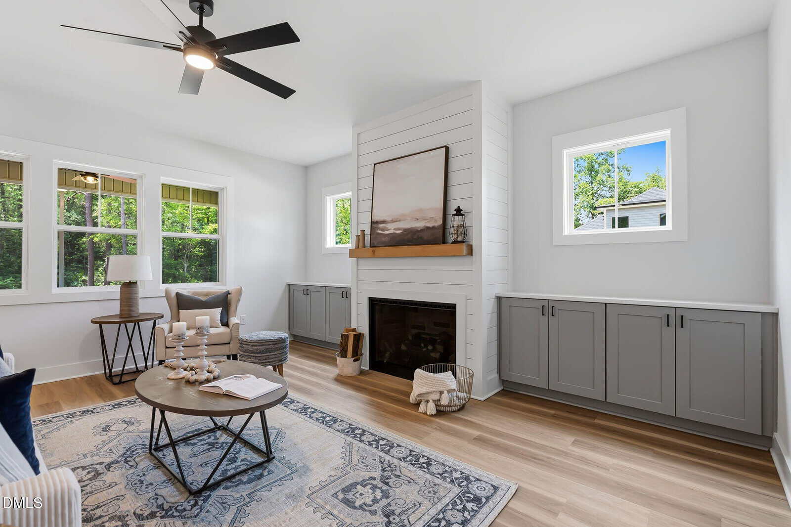 Bright living room with a ceiling fan, fireplace, gray cabinets, large windows, a round coffee table, armchairs, and a blue patterned rug on wood floors. Artwork and decor are placed on the fireplace mantel.