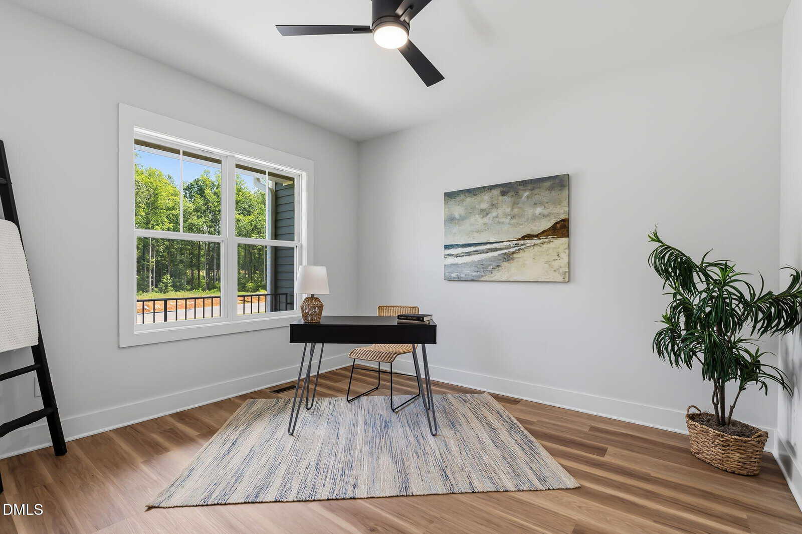 Minimalist home office with a small black desk, chair, lamp, and book on a striped rug. Large window, beach painting on the wall, potted plant, and wood floors create a bright, inviting space.